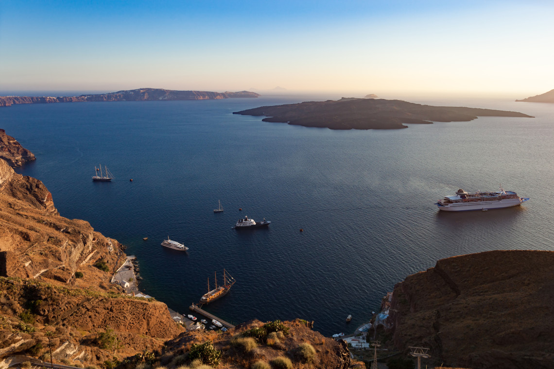 Elevated view across Santorini&rsquo;s caldera in Greece, with cruise ships and sailing boats surrounded by the volcanic islands of the Aegean Sea during the evening light.