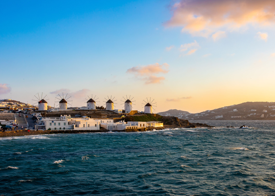 Traditional whitewashed Kato Mili windmills standing beside the coast of Mykonos, Greece, with the Aegean Sea and island hills illuminated by late-day light.