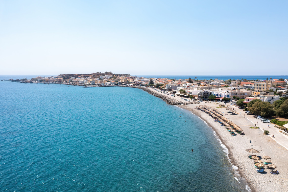 Coastal settlement in Greece extending around a long beach beside clear blue Aegean waters, with houses and waterfront facilities lining the shore.