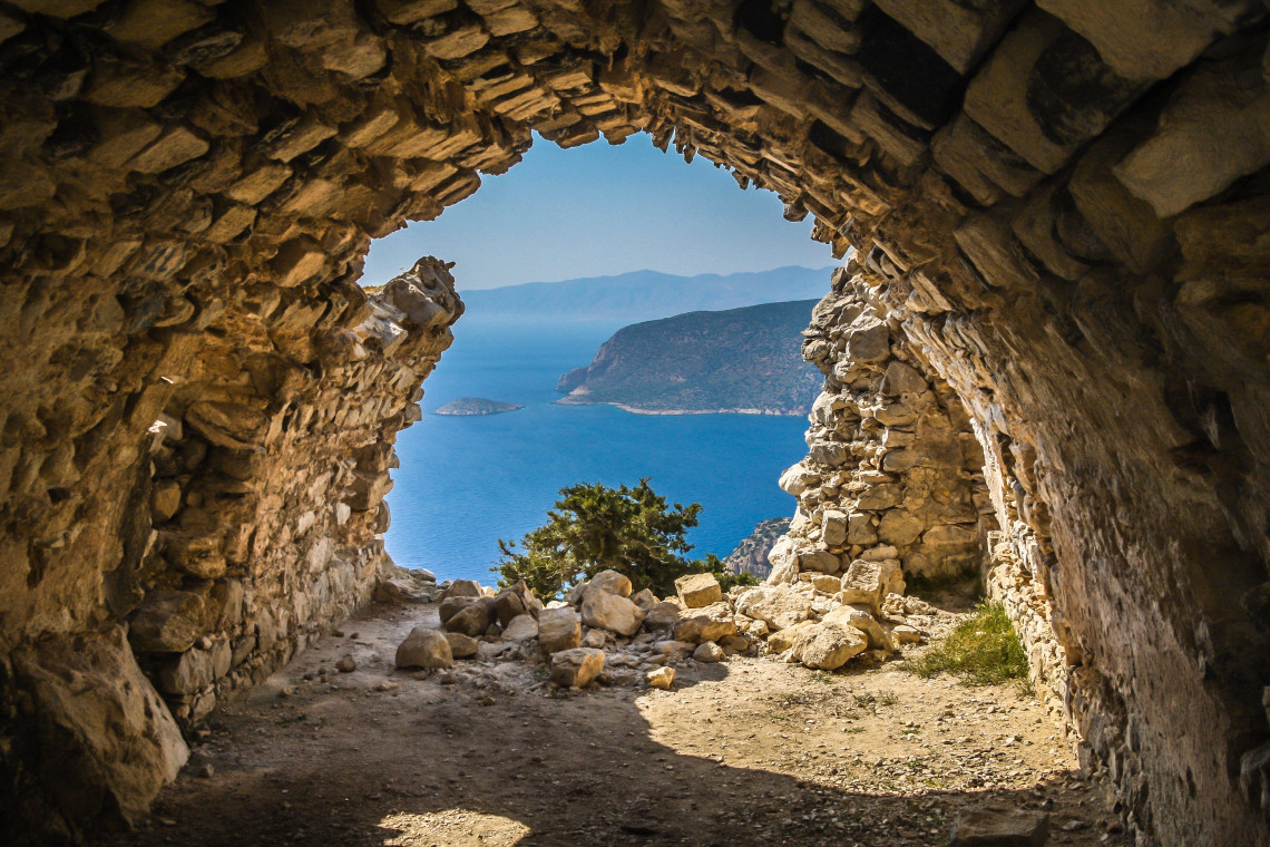 View through the entrance of a rocky coastal cave in the Greek Islands, looking toward steep cliffs, a small offshore islet and the blue waters of the Aegean Sea.