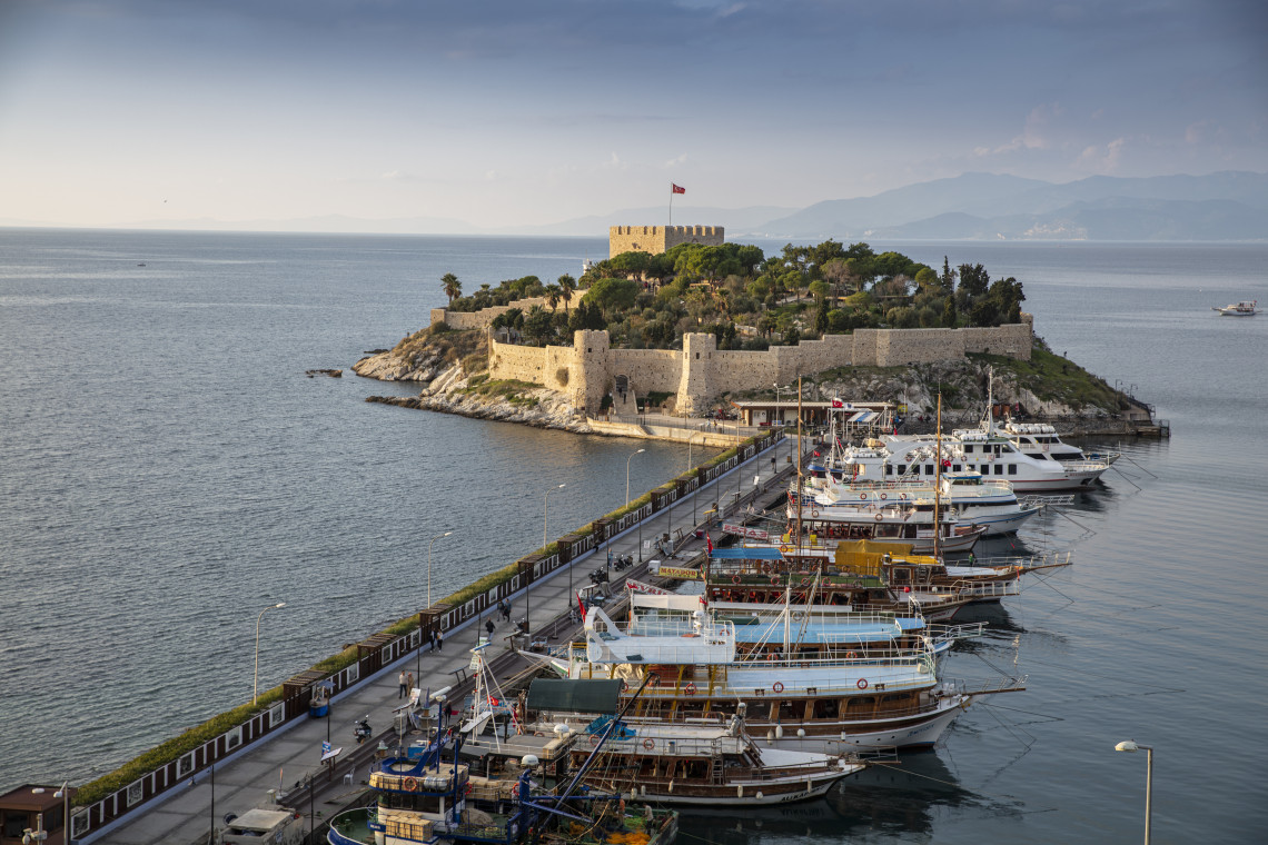 Pigeon Island and its historic stone fortress in Kuşadası, Turkey, connected to the mainland by a causeway beside a harbour filled with excursion boats.