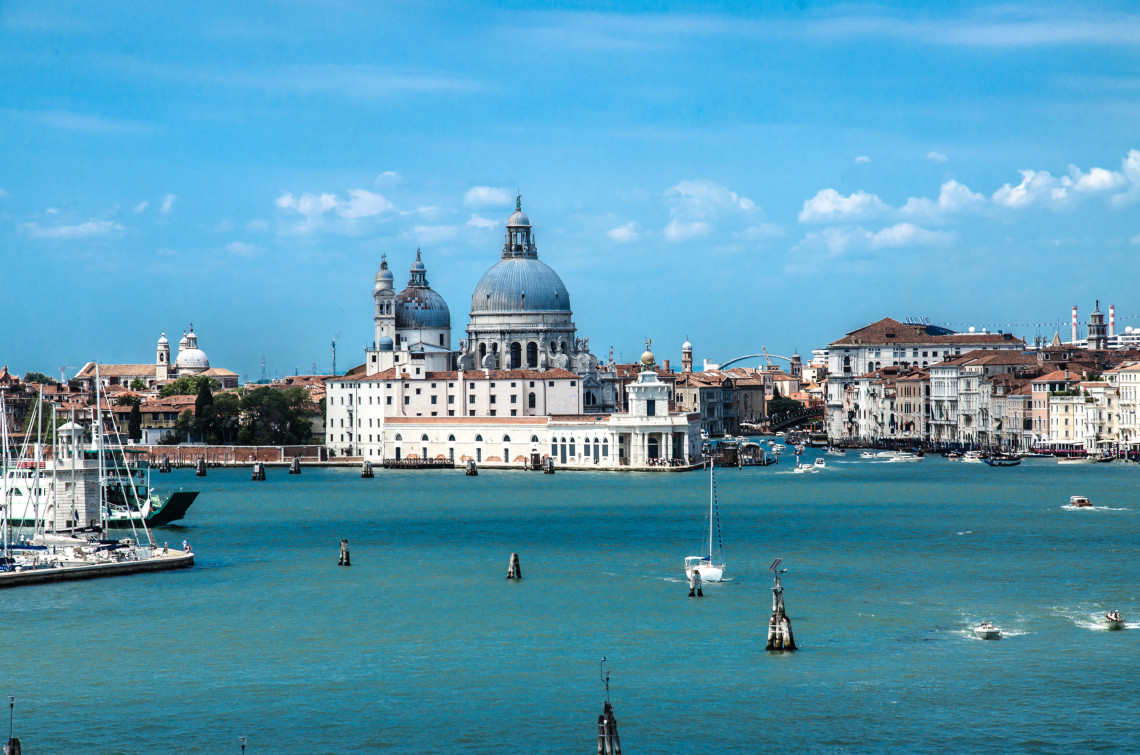 Basilica di Santa Maria della Salute rising above the waterfront in Venice, Italy, with boats crossing the Grand Canal and historic buildings extending along the shoreline.