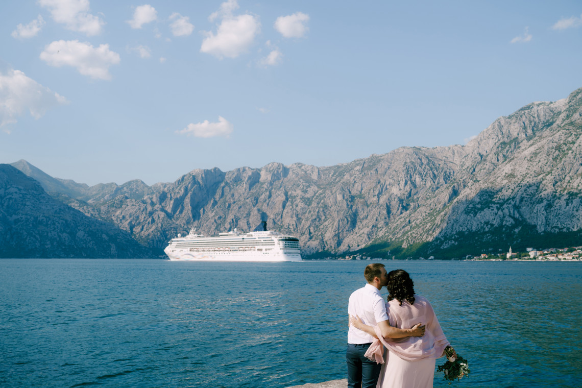 Cruise ship sailing through the Bay of Kotor in Montenegro beneath steep limestone mountains, while a couple looks across the calm Adriatic waters from the waterfront.