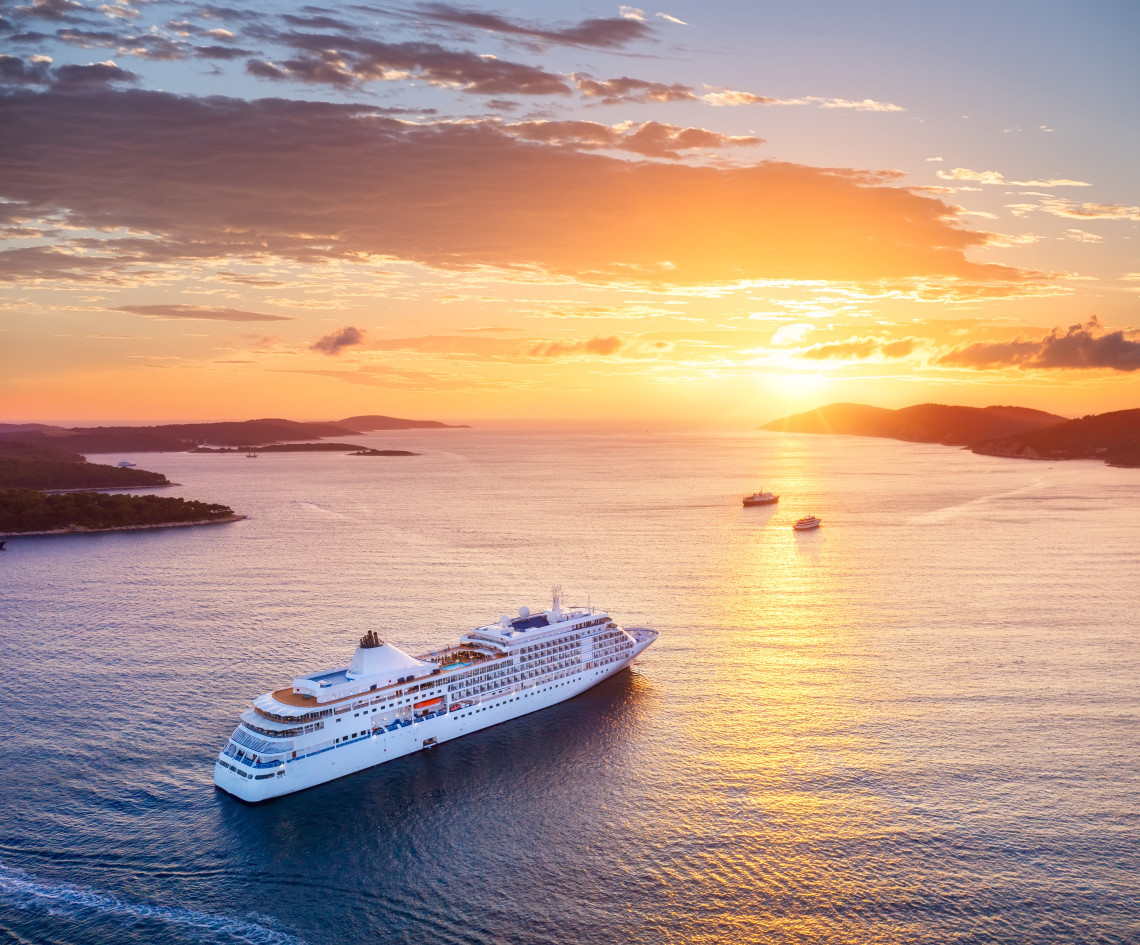 Cruise ship sailing through the Adriatic Sea at sunset, surrounded by low islands and distant vessels beneath a warm orange sky.