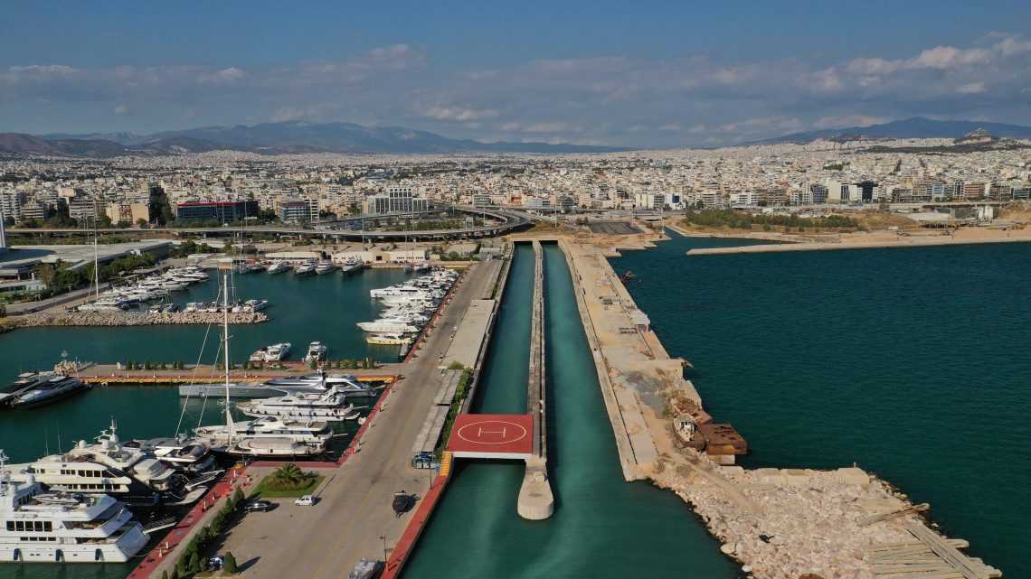 Aerial view of a marina along the Athens Riviera in Greece, with yachts moored beside long piers and the dense urban landscape of Athens extending inland.