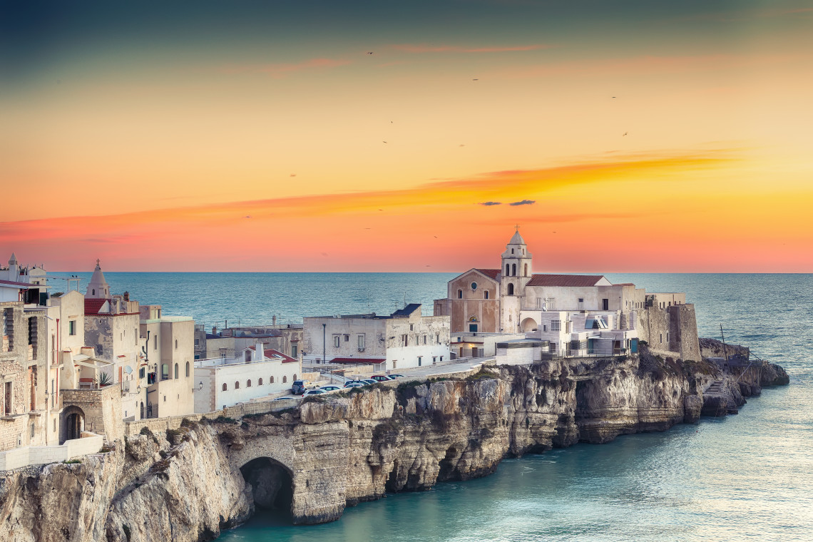 Historic centre of Vieste in Puglia, Italy, built along limestone cliffs above the Adriatic Sea, with churches and stone buildings illuminated by the colours of sunset.