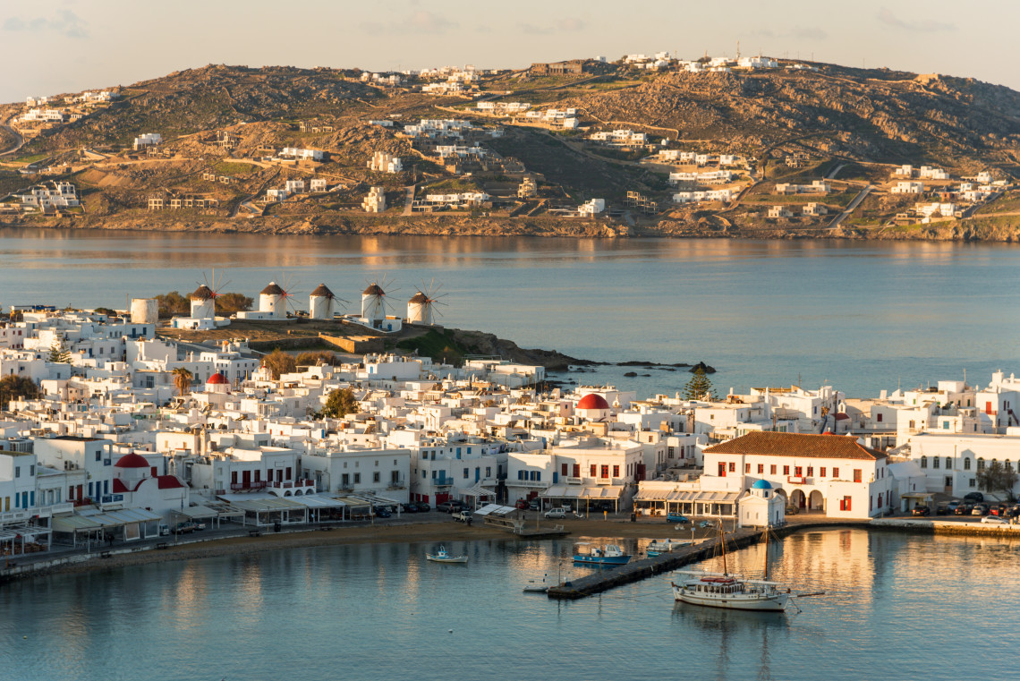 Panoramic view of Mykonos Town in Greece, with whitewashed buildings surrounding the harbour and the Kato Mili windmills standing on the coastline beyond.