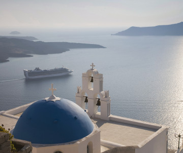 Blue-domed church overlooking the Santorini caldera in Greece, with a cruise ship sailing through the Aegean Sea and volcanic islands visible in the distance.