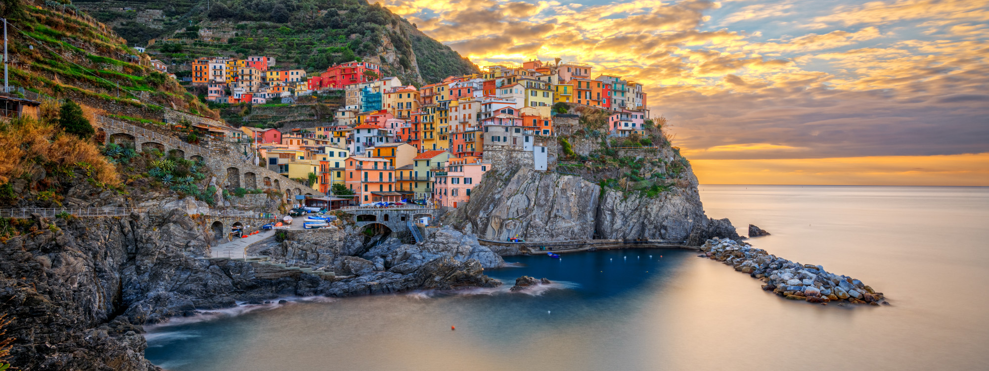 Manarola rises above a small rocky harbour on the Cinque Terre coast in Liguria, Italy. Colourful houses cluster on steep cliffs above the Mediterranean as evening light spreads across the sea.