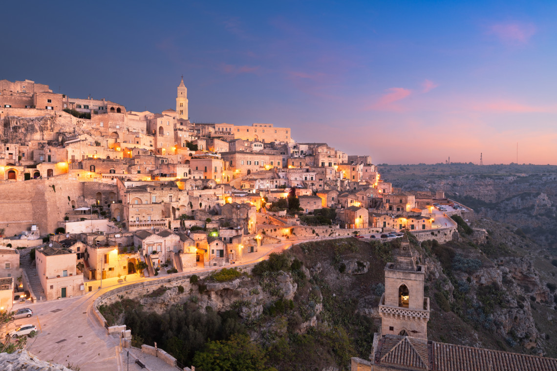 The Sassi di Matera spread across the rocky slopes of Matera in Basilicata, Italy, as evening lights illuminate stone houses and cave dwellings. Church towers rise above the layered historic city beside the surrounding ravine.