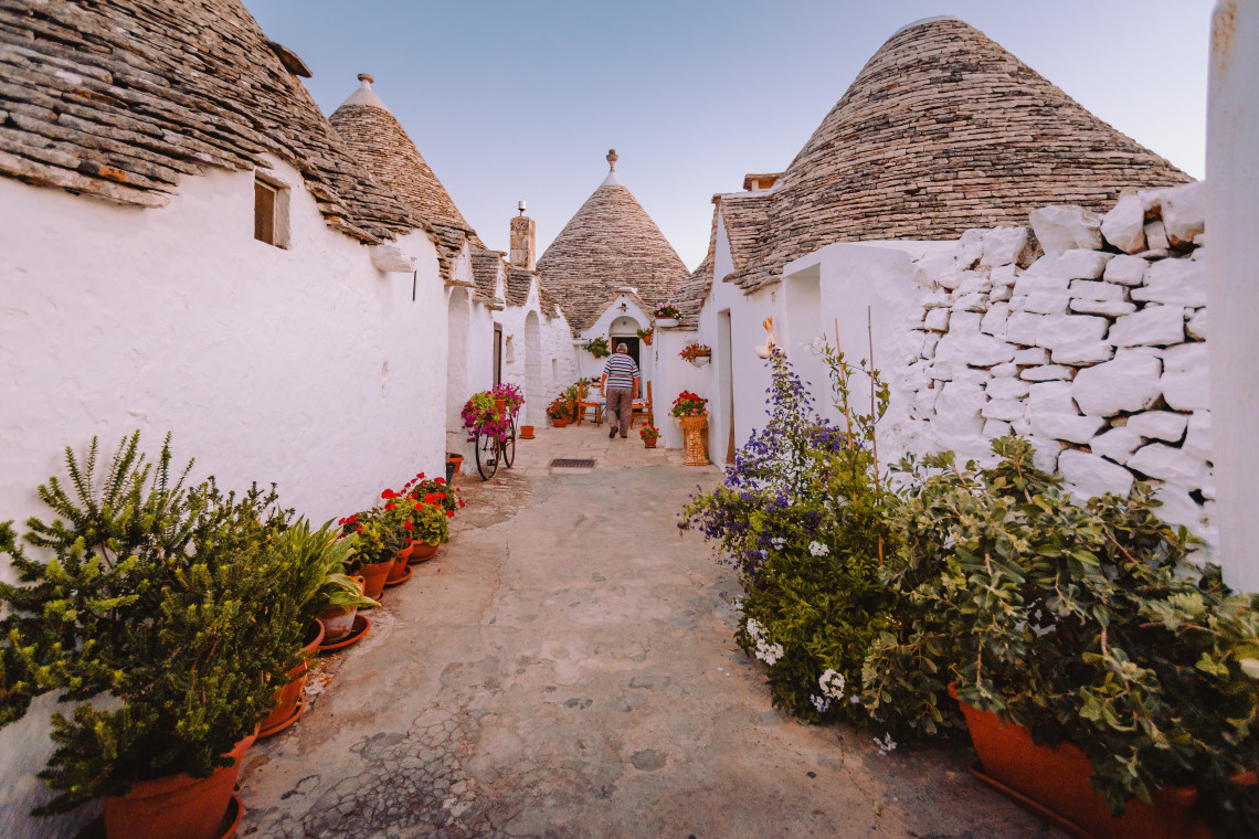 Traditional trulli line a narrow lane in Alberobello, Puglia, Italy, with whitewashed stone walls and distinctive conical roofs. Potted flowers and plants decorate the pedestrian street between the historic houses.