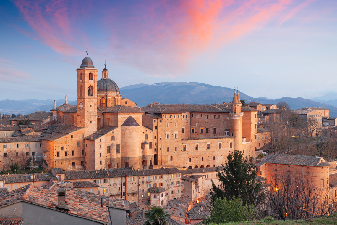 The historic centre of Urbino in the Marche region of Italy is illuminated by warm evening light. The Renaissance cityscape includes the monumental Palazzo Ducale complex surrounded by terracotta rooftops and rolling hills.