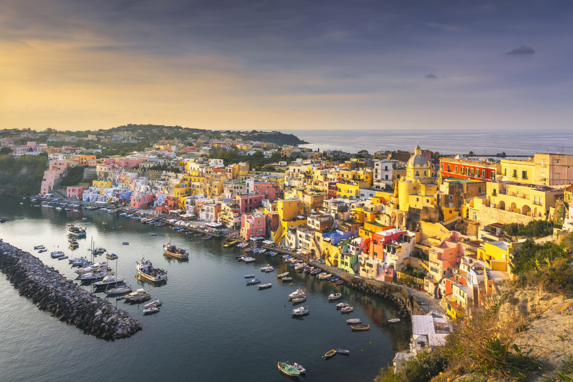 Marina Corricella curves around a sheltered harbour on Procida Island in Campania, Italy. Pastel-coloured waterfront houses, fishing boats and the domed church create the distinctive coastal landscape.