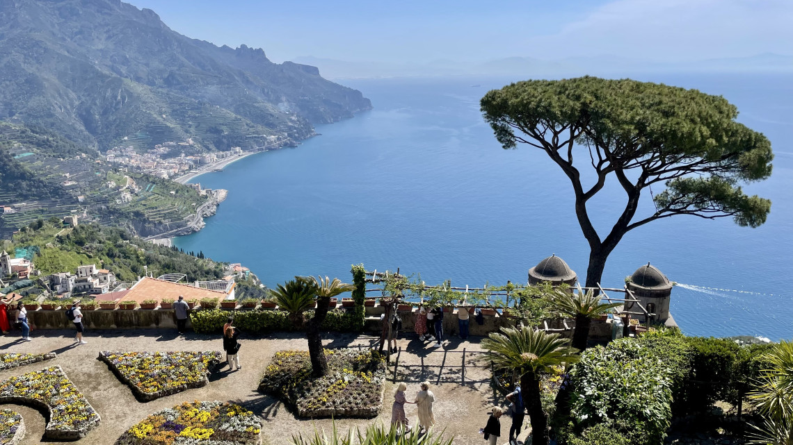 The gardens of Villa Rufolo in Ravello overlook the Amalfi Coast and Mediterranean Sea in Campania, Italy. A tall umbrella pine, landscaped flower beds and historic garden structures frame the coastal panorama.