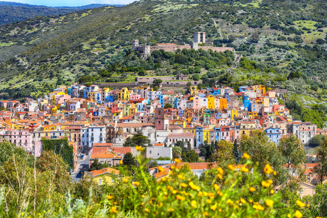 Colourful houses cover the hillside of Bosa in Sardinia, Italy, beneath the medieval Malaspina Castle. Green Mediterranean vegetation surrounds the tightly packed historic neighbourhood.