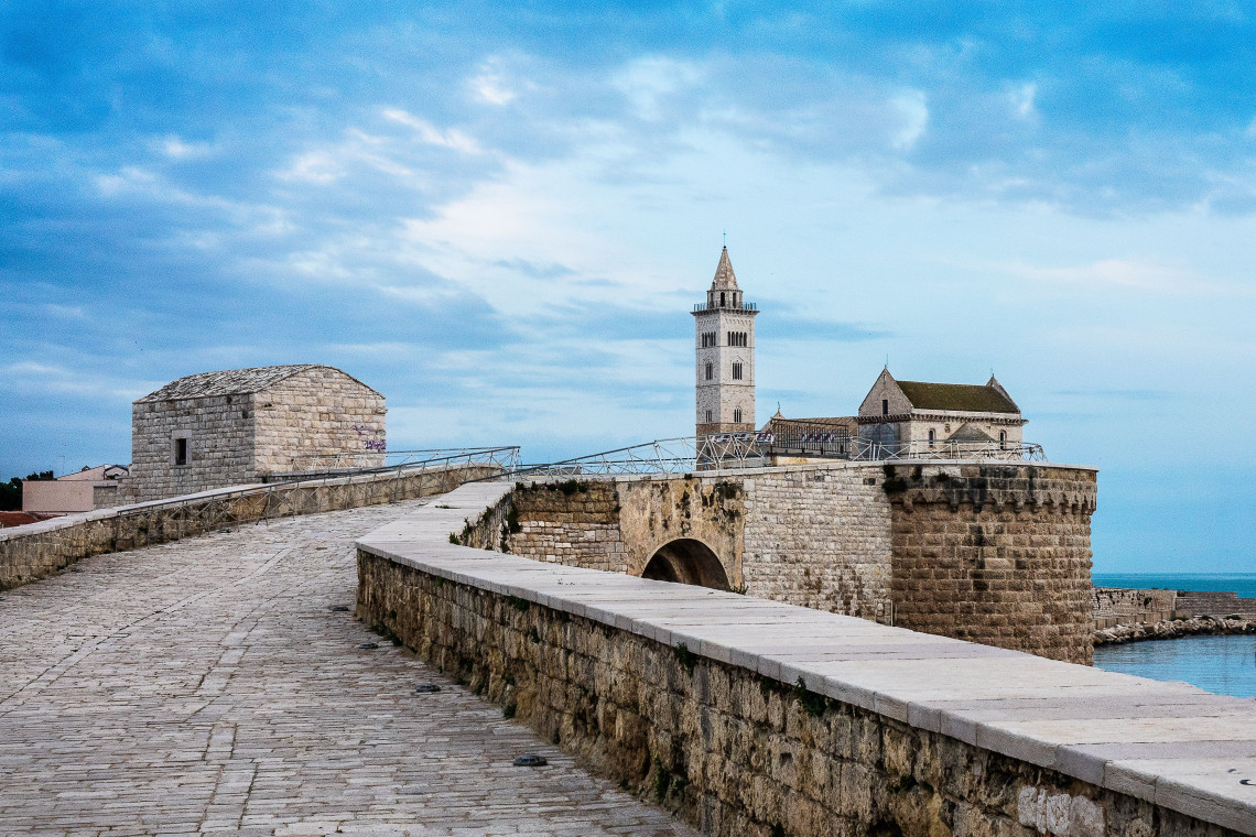 Trani Cathedral stands beside the Adriatic Sea in Trani, Puglia, Italy, with its tall bell tower and pale stone exterior rising above the fortified waterfront. A broad stone walkway leads toward the cathedral.