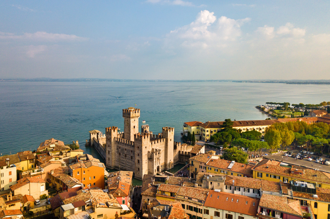 Scaliger Castle rises above the historic centre of Sirmione on Lake Garda in Italy. Crenellated towers and fortified walls stand beside the lake, surrounded by terracotta-roofed buildings.