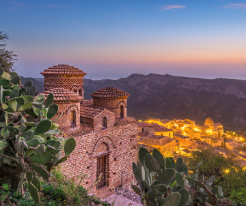 The Cattolica di Stilo overlooks the historic village of Stilo in Calabria, Italy, at dusk. Byzantine brickwork, tiled domes and prickly pear cacti frame the church against the surrounding mountain landscape.