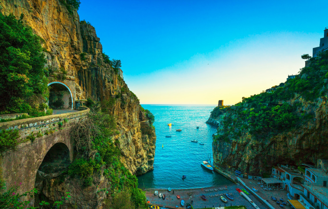 Dramatic view of Furore Fjord with a tunnel road carved into limestone cliffs, a hidden pebble beach at the bottom and boats on the turquoise Mediterranean Sea, Amalfi Coast, Campania, Italy