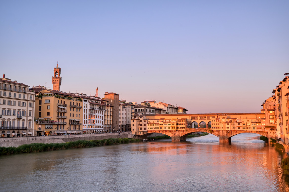 Ponte Vecchio medieval bridge reflected in the Arno River at sunset with Palazzo Vecchio tower visible in the Florence skyline, Tuscany, Italy