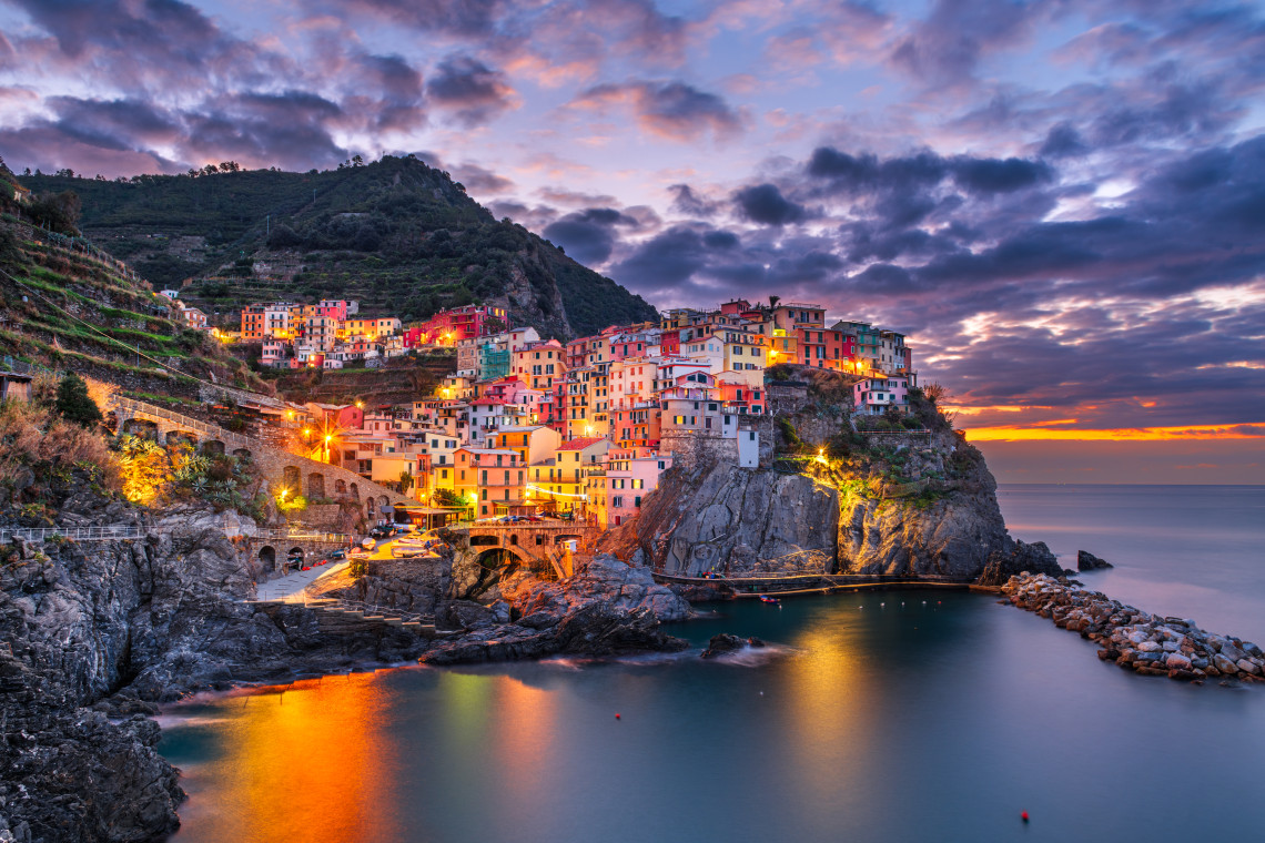 Dramatic dusk view of Manarola village with illuminated colorful buildings perched on rocky cliffs above the Ligurian Sea, Cinque Terre, Liguria, Italy