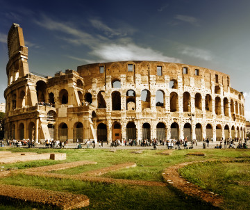 Exterior view of the Colosseum ancient Roman amphitheatre with tourists and grass in the foreground under a dramatic cloudy sky, Rome, Italy