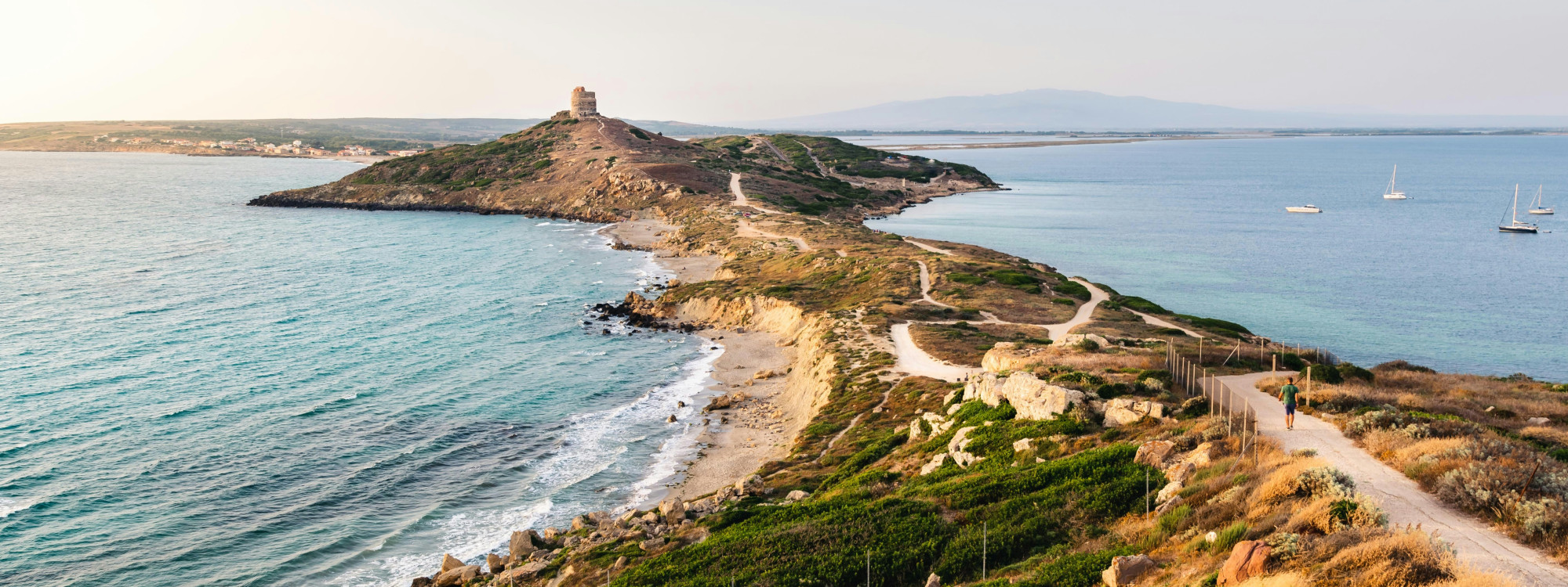 Aerial view of Torre di San Giovanni ancient watchtower on a rocky headland with turquoise Mediterranean waters and a winding coastal road, Sardinia, Italy