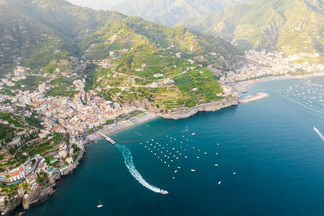 Wide aerial view of Amalfi town with terraced lemon groves cascading down steep mountains to the beach and marina with boats on the deep blue Mediterranean Sea, Campania, Italy