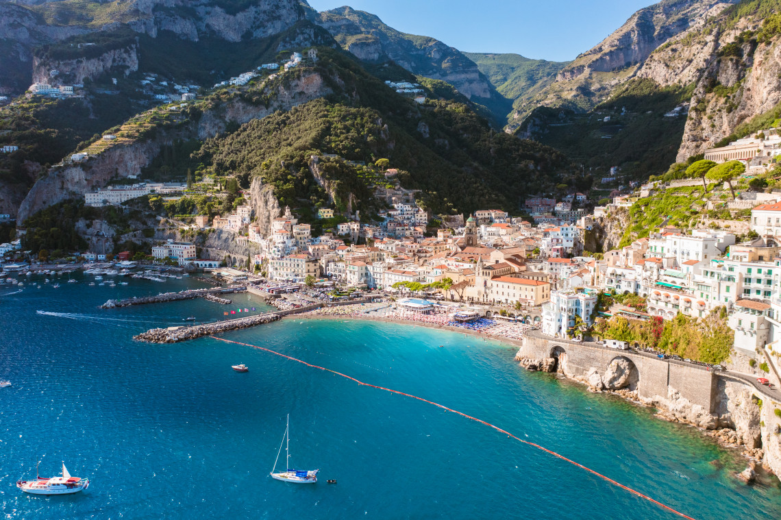 Aerial view of Amalfi town with crowded beach, colorful buildings, cathedral and sailboats in the turquoise bay surrounded by dramatic limestone cliffs, Campania, Italy