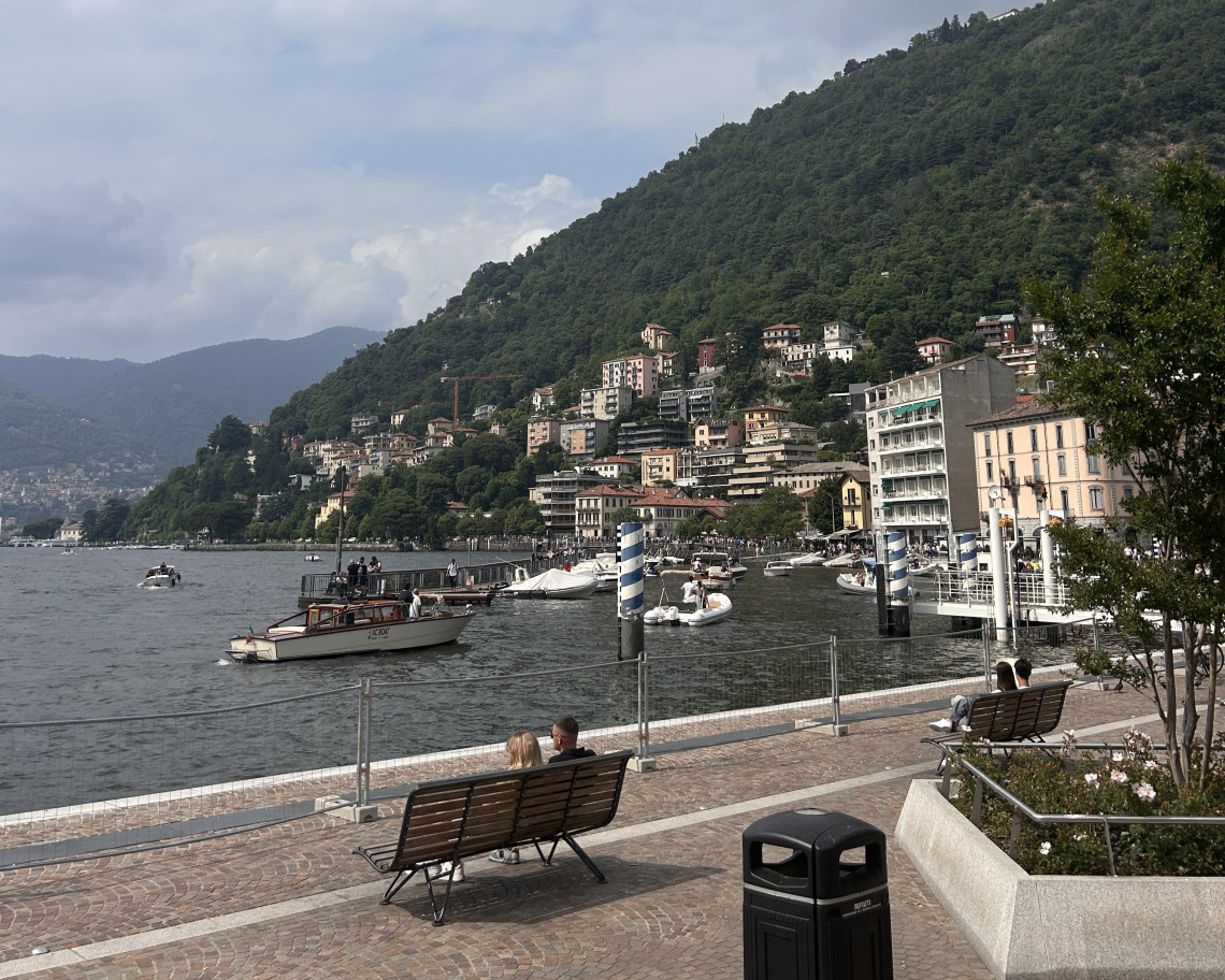 Couple sitting on a bench along the Como town lakeside promenade with ferry boats, waterfront buildings and forested Alpine hills, Lake Como, Lombardy, Italy