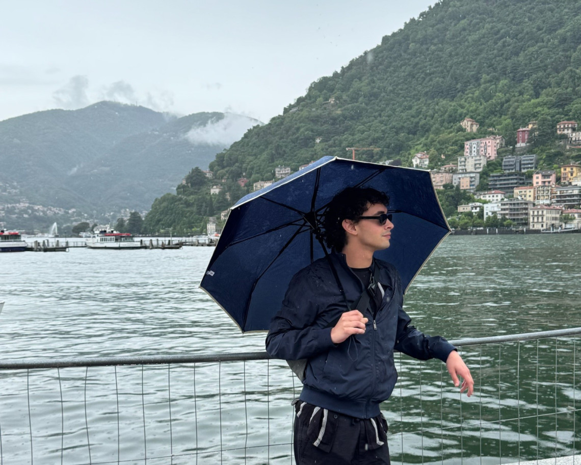 Young male traveler holding an umbrella on the Como town waterfront on a rainy day with colorful lakeside villages and forested hills across the lake, Lake Como, Lombardy, Italy