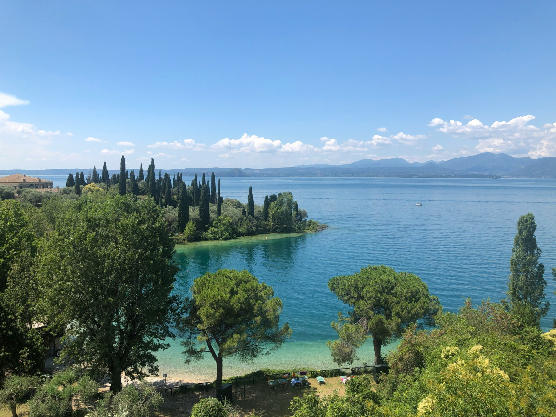 Panoramic view of Punta San Vigilio cypress tree peninsula jutting into the turquoise waters of Lake Garda with the Alpine foothills in the distance, Veneto, Italy