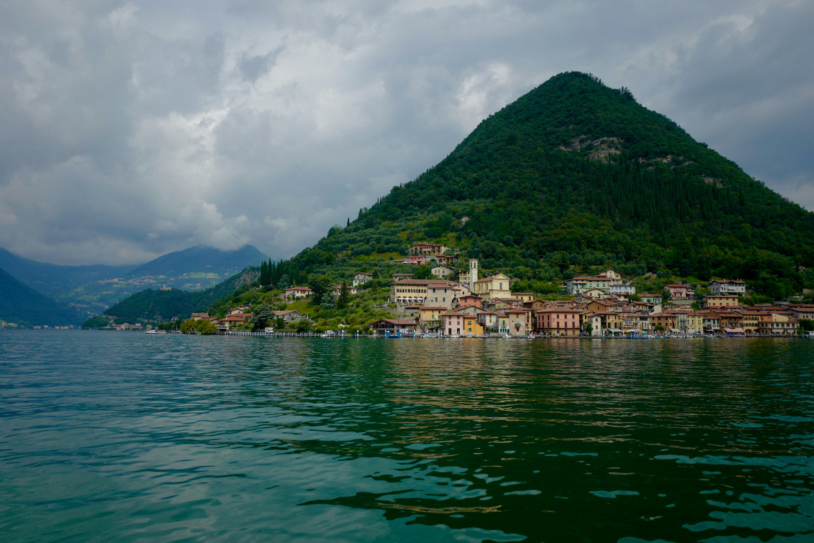 View from the water of Monte Isola lakeside village with colorful buildings at the base of a forested conical hill reflected in the turquoise waters of Lake Iseo, Lombardy, Italy
