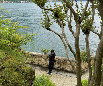 Traveler walking along a scenic lakeside path with ornate stone balustrade overlooking the turquoise waters of Lake Como surrounded by lush vegetation, Lombardy, Italy