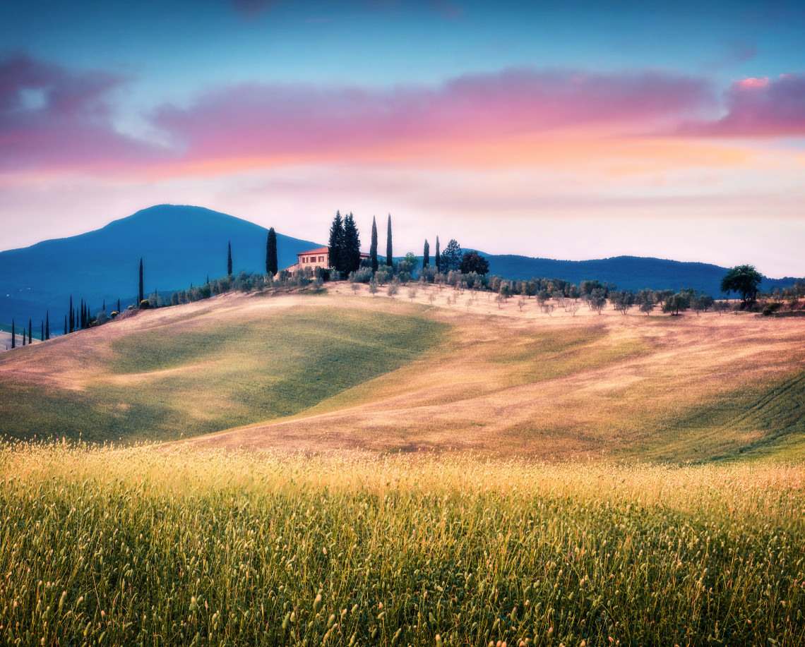 Rolling fields in Val d&rsquo;Orcia, Tuscany, surround a rural farmhouse framed by tall cypress trees, with soft hills and mountains in the background.