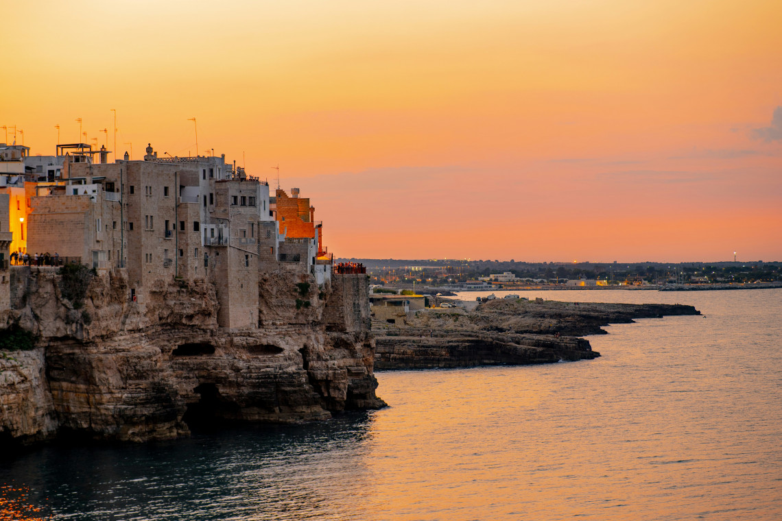 Historic buildings of Polignano a Mare rise directly above limestone cliffs on the Adriatic coast of Puglia, Italy, illuminated by the warm colours of sunset.