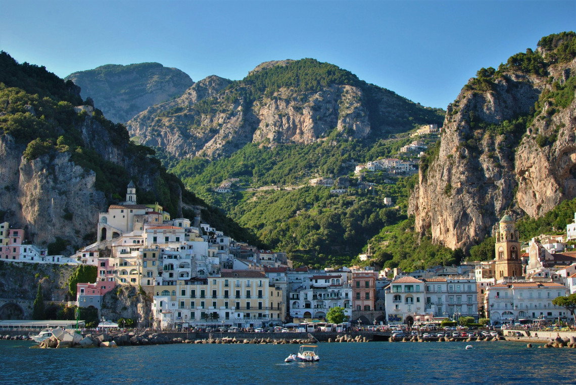 Waterfront view of Amalfi on Italy&rsquo;s Amalfi Coast, with colourful historic buildings clustered beneath steep green mountains and rocky cliffs beside the Tyrrhenian Sea.