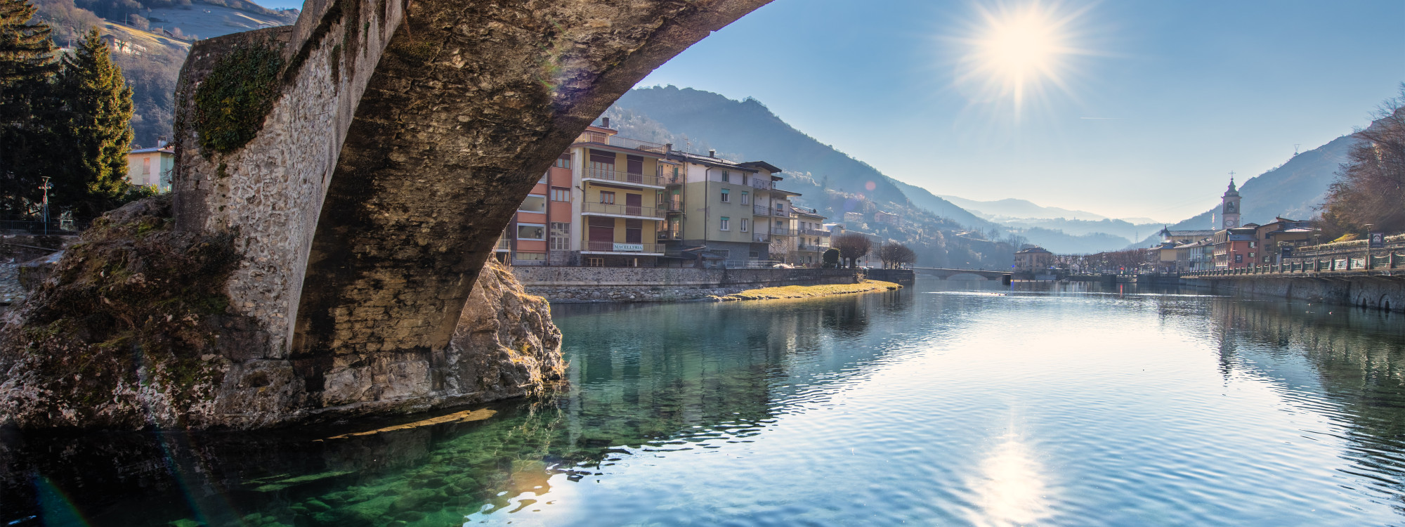 View from under the historic Ponte San Nicola stone arch bridge over the clear Brembo River with the Alpine town of Bergamo in the background, Lombardy, Italy