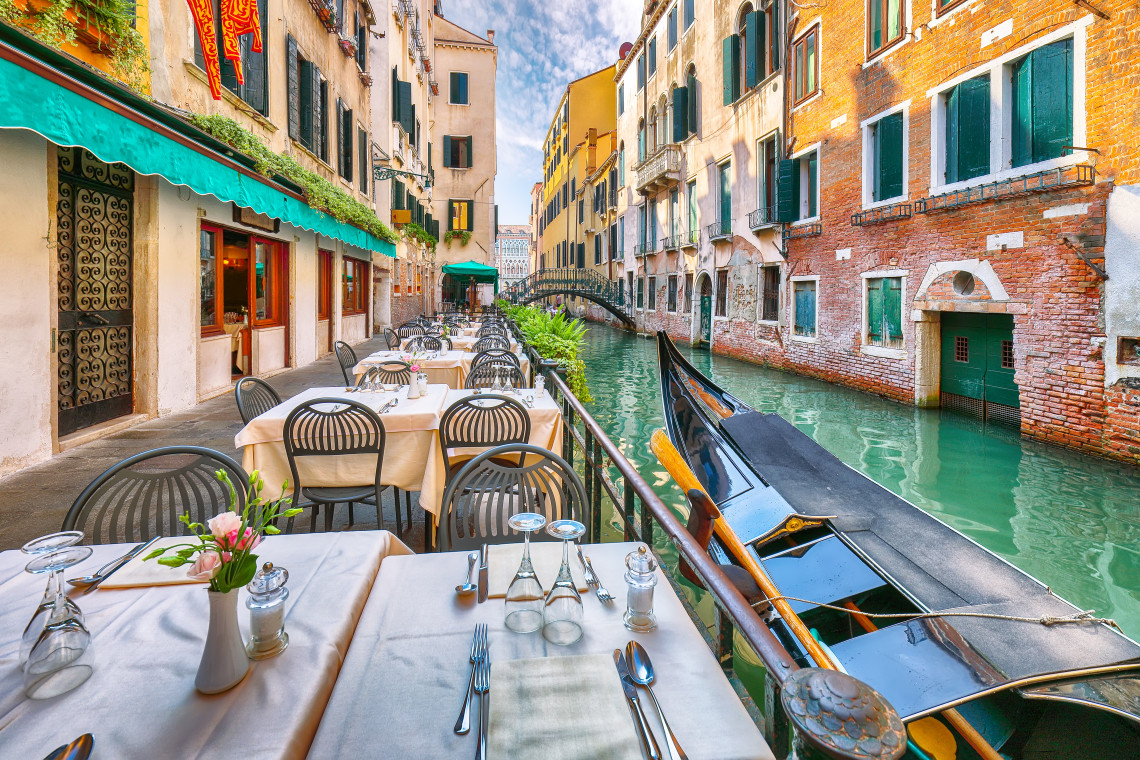 Outdoor restaurant tables set along a narrow Venice canal with a gondola moored alongside historic brick buildings, Venice, Italy