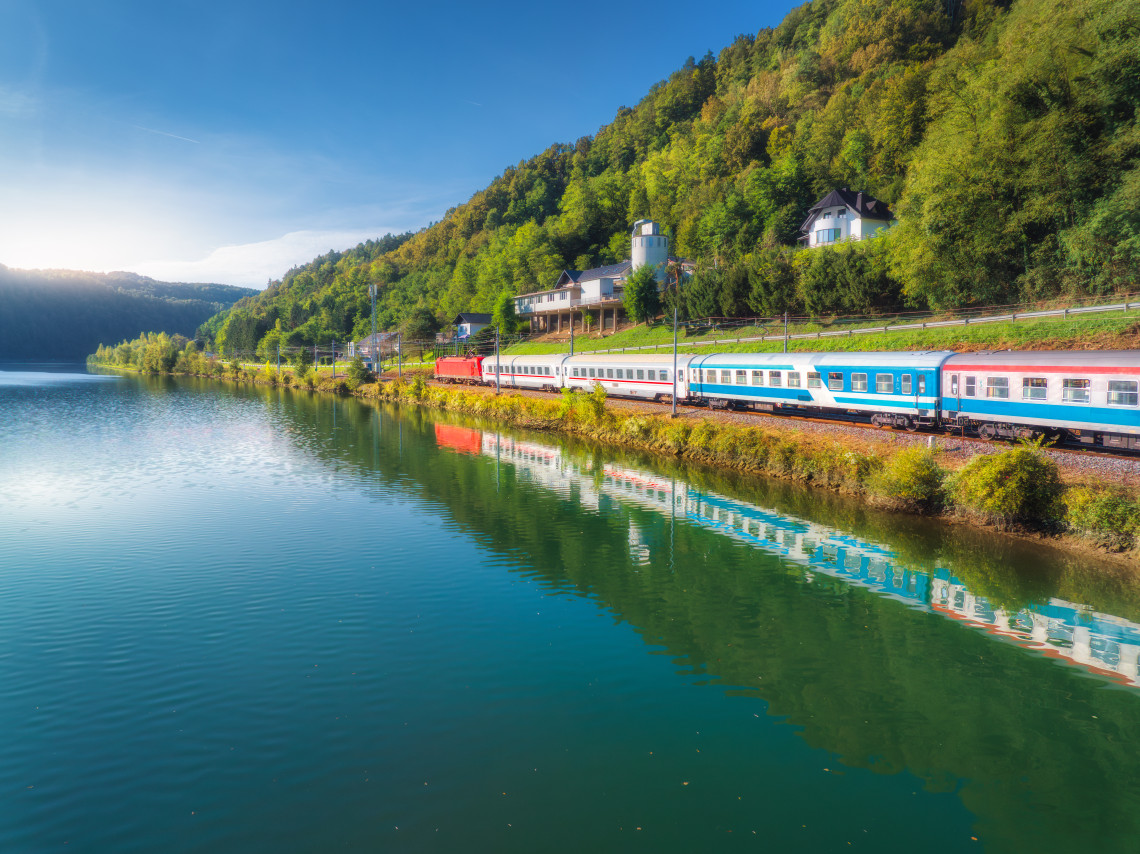 Blue and red passenger train traveling alongside a calm mountain lake with forested hills reflected in the water, Italy