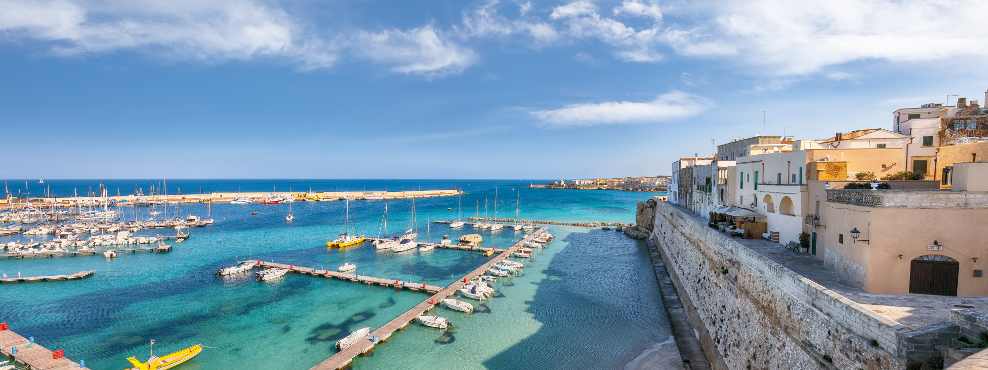 View across the Port of Otranto in Puglia, with boats moored in turquoise Adriatic waters beside the stone walls and buildings of the historic waterfront.