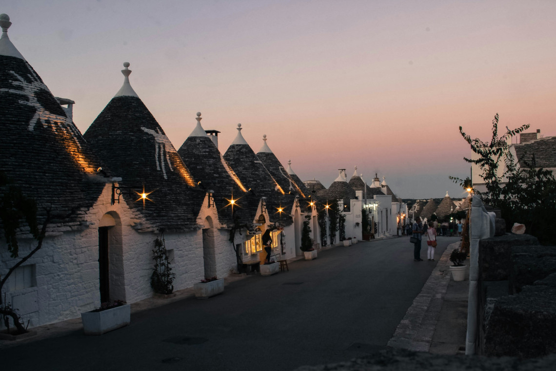 Traditional whitewashed trulli with conical stone roofs lining a street in Alberobello, Puglia, photographed as evening lights begin to illuminate the historic district.
