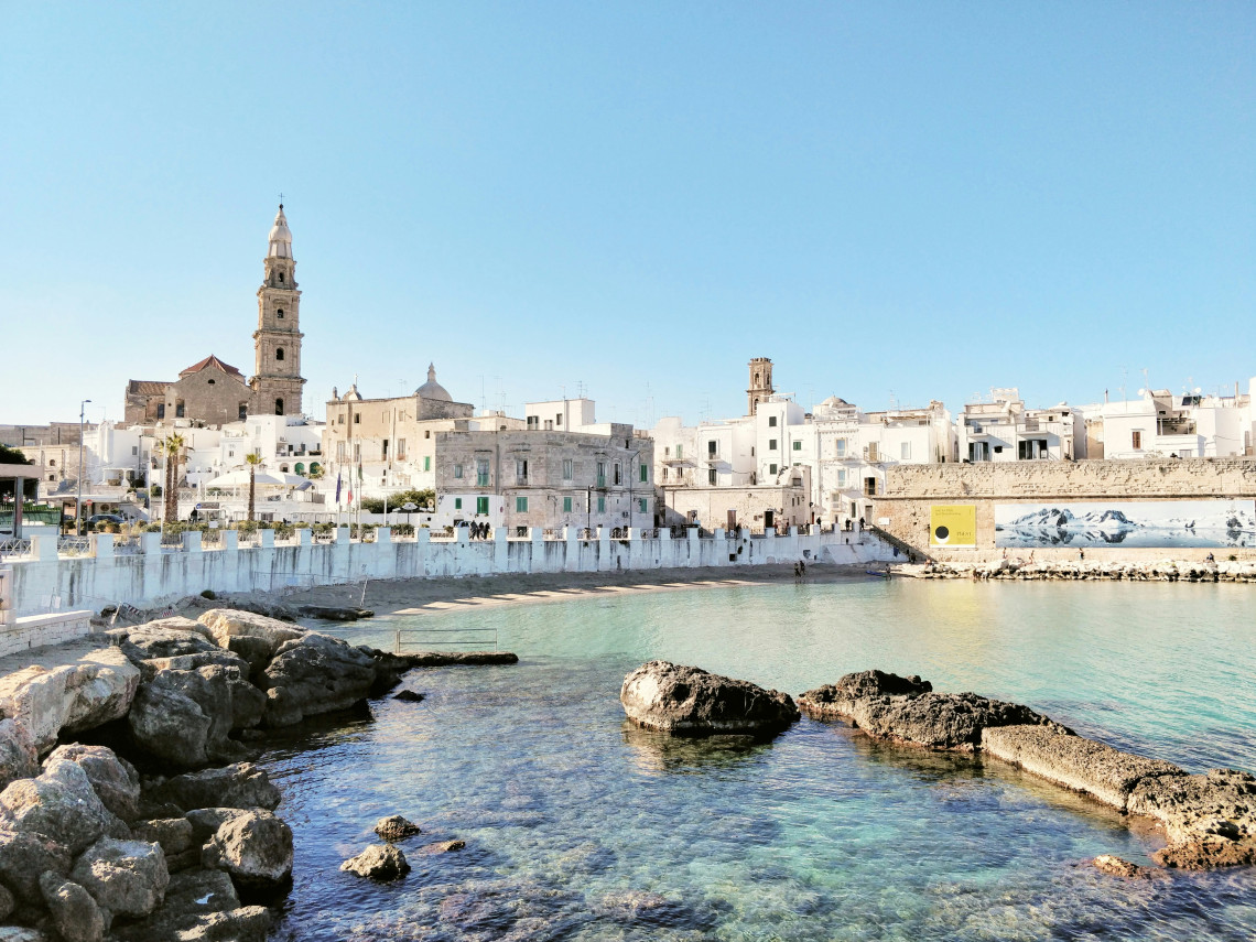 Cala Porta Vecchia in Monopoli, Puglia, with clear Adriatic water, rocky shoreline and the white buildings and church towers of the historic centre behind the beach.