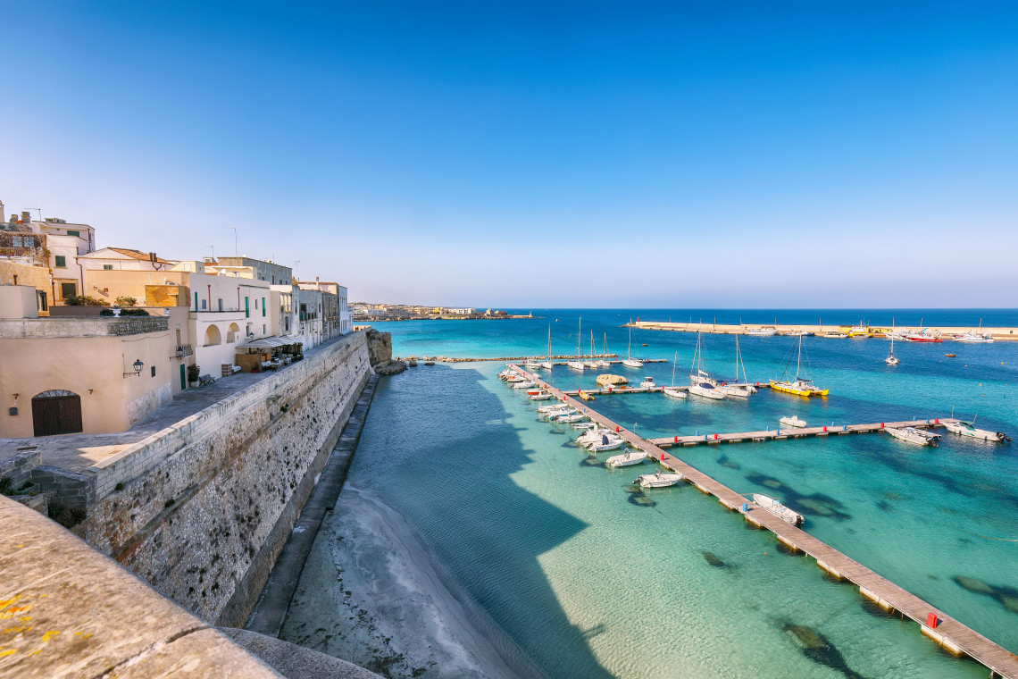 Port of Otranto in Puglia viewed from the historic waterfront, with sailboats and small vessels moored in clear turquoise water beside the old city walls.