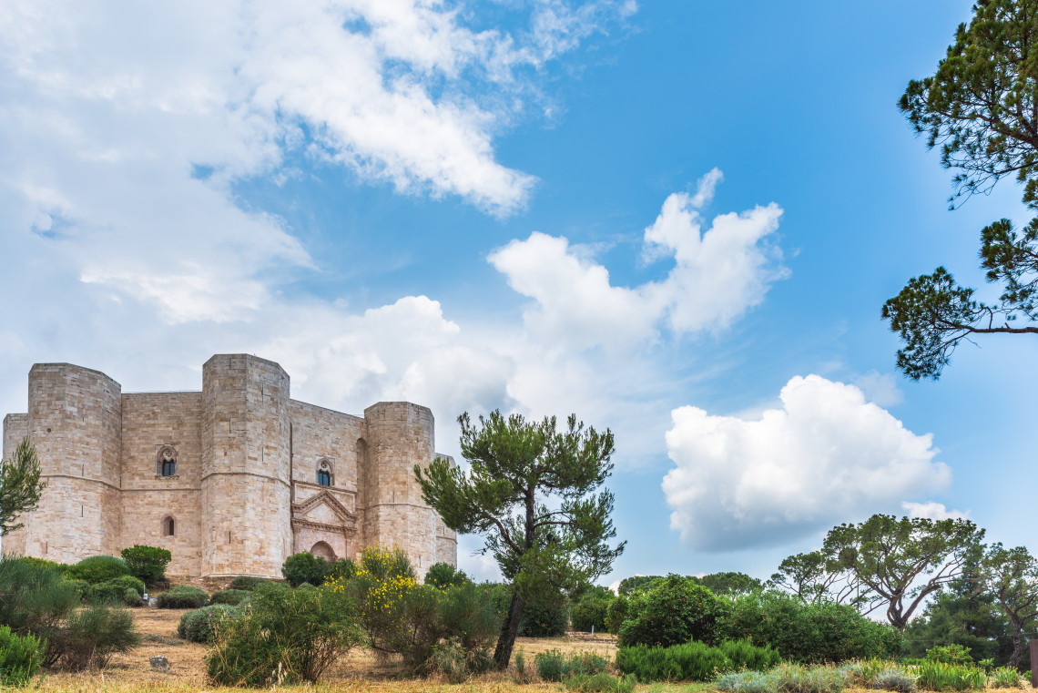 Castel del Monte near Andria, Puglia, surrounded by Mediterranean vegetation beneath a blue sky. The medieval stone fortress is distinguished by its octagonal structure and towers.
