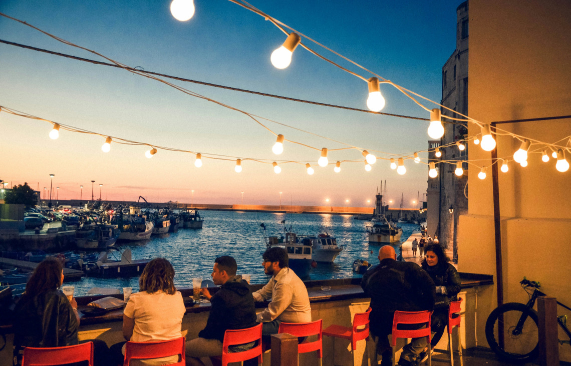 Evening scene at Porto Vecchio in Monopoli, Puglia, where people sit along the harbourfront beneath hanging lights as fishing boats rest in the marina at sunset.