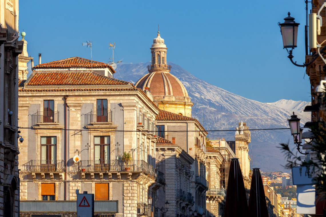 Baroque street in Catania with ornate church dome and snow capped Mount Etna visible above the city rooftops, Sicily, Italy