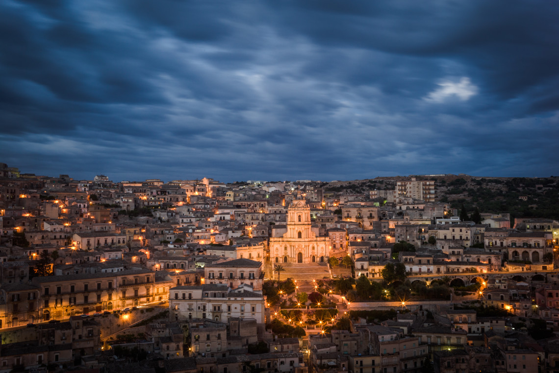 Panoramic night view of Modica with illuminated Baroque buildings and San Giorgio Cathedral glowing against a dramatic cloudy sky, Sicily, Italy