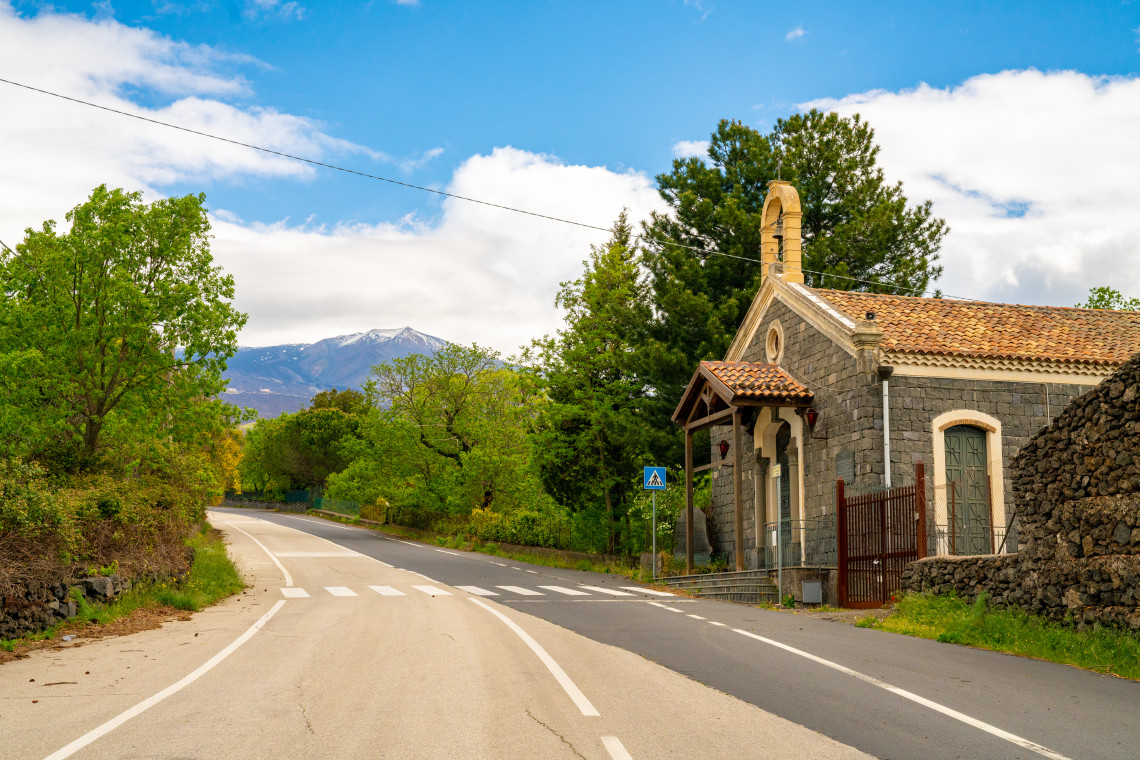 Empty rural road through the Etna foothills with a traditional lava stone chapel and snow capped Mount Etna visible in the background, Sicily, Italy