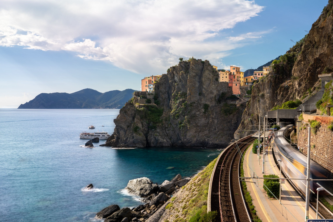 Manarola village perched dramatically on rocky cliffs above the Cinque Terre coastal railway station with a ferry boat and the Ligurian Sea in the background, Liguria, Italy
